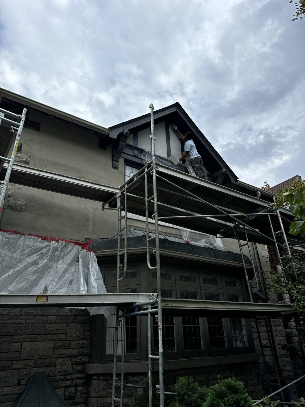 Stucco being applied by hand to a Tudor gable from scaffolding, with windows masked off